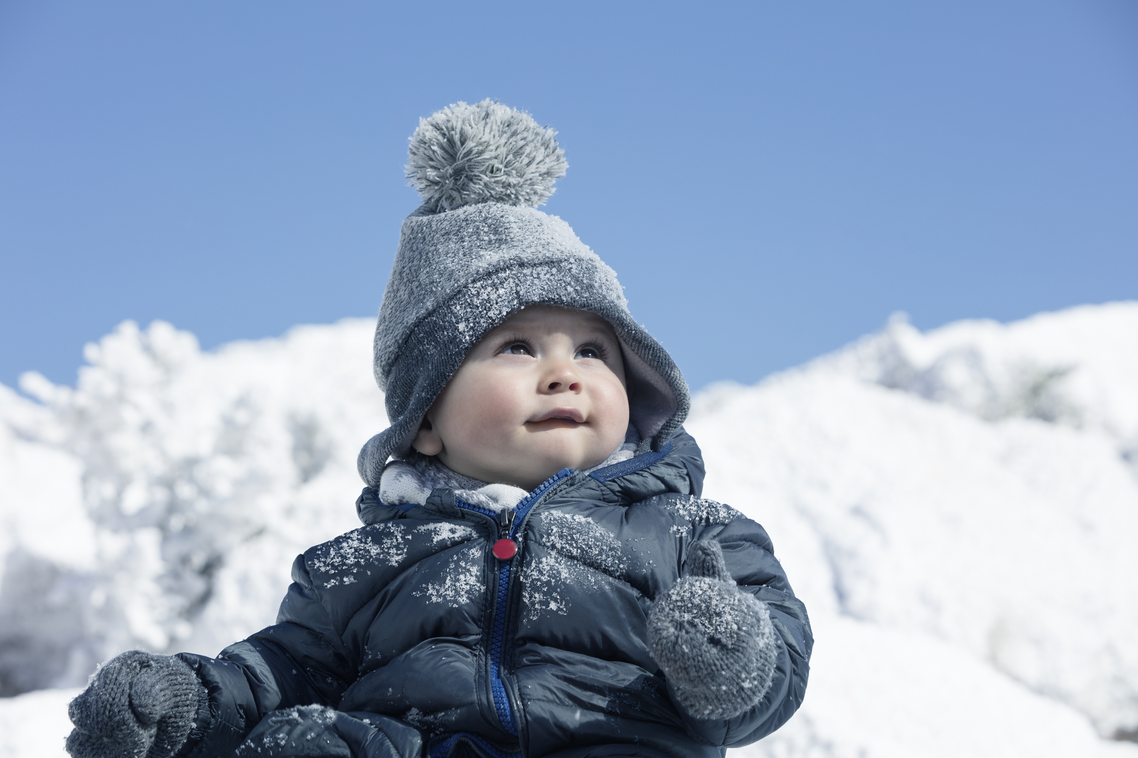 Baby dressed in a thick winter jacket, knitted hat with a pom-pom, and gloves, sitting outdoors surrounded by bright white snow under a clear blue sky.